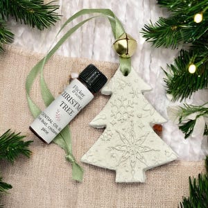 May include: A festive image showing a white Christmas tree ornament with snowflake patterns, a small essential oil bottle, and a gold bell. The bottle is labelled "Christmas Tree" with essential oil ingredients. A green ribbon is attached to the ornament and bell.