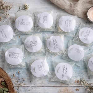 May include: An overhead shot of several small, round, white products in clear packaging, each labeled with text. The labels include scent names like "Black Ne & Honey" and "Wood Sage & Sea Salt." A lit candle and dried flowers are also present.