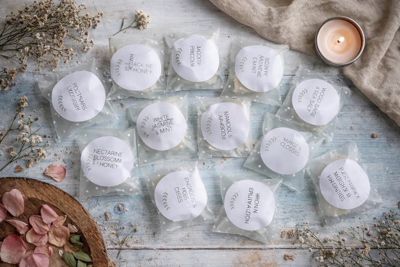 May include: An overhead shot of several small, round, white products in clear packaging, each labeled with text. The labels include scent names like "Black Ne & Honey" and "Wood Sage & Sea Salt." A lit candle and dried flowers are also present.