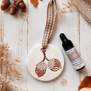 May include: A round white clay ornament featuring a stamped brown acorn design, suspended by a brown and white gingham ribbon, accompanied by a 5ml amber glass dropper bottle labelled "Tucker & Scent calm ESSENTIAL OIL BLEND".