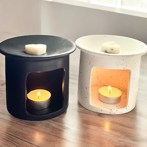 May include: Two ceramic wax warmers, one black and one white with speckles. Both have a heart-shaped wax melt on top and a tea light candle in the bottom.