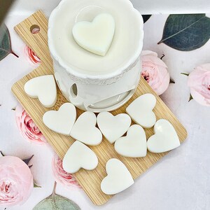 May include: A white ceramic wax warmer with a heart-shaped wax melt on top, and several heart-shaped wax melts on a small wooden board. The warmer and melts are white, and the board is light brown. The background has a floral pattern.