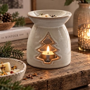 May include: A cream-coloured ceramic wax warmer featuring a cutout in the shape of a pine tree. The base houses a lit tea light candle, while the top bowl holds wax melts. The glaze has a smooth, glossy finish with subtle brown edges.