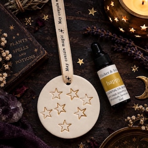 May include: A round white ceramic ornament with gold-coloured star engravings, suspended by a cream ribbon printed with the text May contain magic. Beside it sits a 5ml amber glass dropper bottle labelled Flicker & Soul happy essential oil blend.