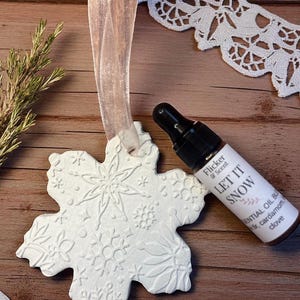 May include: A white snowflake-shaped ornament with embossed snowflake designs, suspended by a sheer ribbon. A small essential oil bottle with the text "LET IT SNOW" is next to the ornament. The bottle has a black dropper top.
