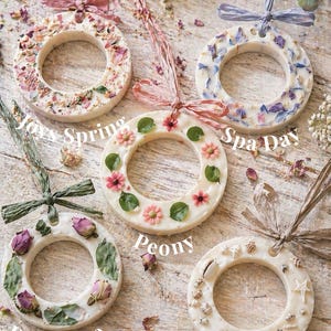 May include: Five circular wax melts decorated with dried flowers, petals, and seashells. Each melt has a ribbon for hanging. The melts are labelled with the text: "Joy's Spring", "Spa Day", "Peony", "Romance", and "Coastal Walk".
