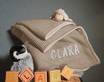 May include: A beige waffle weave baby blanket with the name "CLARA" embroidered on it. The blanket is folded and displayed with a stuffed penguin and a stuffed lamb, and wooden blocks that spell out the name "CLARA".