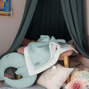 May include: A teal waffle weave blanket with a white sherpa lining. The blanket is folded and draped over a small wooden stool with a pink cushion. There are several pillows in the background, including a large green crescent-shaped pillow with a white embroidered design.