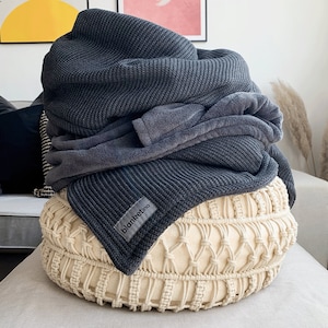 May include: A dark grey knitted blanket and a lighter grey fleece blanket are arranged on a cream macrame pouffe. The dark grey blanket has a label that reads "blanketino". The setting is a bright room with artwork on the wall.