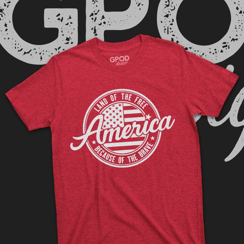 May include: Red t-shirt with a white circle graphic featuring a stylized American flag and the text "Land of the Free America Because of the Brave".