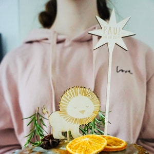 Könnte beinhalten: Ein gefrosteter Kuchen mit blauem Zuckerguss, verziert mit einem hölzernen Löwen und einem Sternentopper. Der Sternentopper trägt den Namen "PAUL" und das Datum "08.08.2019". Der Kuchen befindet sich auf einem blauen Teller mit einem Zweig Rosmarin und Sternanis.