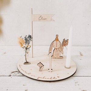 May include: Wooden first birthday cake topper with a bunny wearing a party hat, a candle, a flag with the name "Elise" and the text "du wirst so geliebt" (you are so loved).