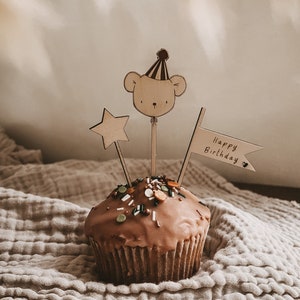 Könnte beinhalten: Ein Schokoladen-Cupcake mit Streuseln und einem hölzernen Bären-Topper, der einen Partyhut trägt. Der Cupcake befindet sich auf einer beigefarbenen, strukturierten Oberfläche mit einer Holzflagge, auf der "Happy Birthday" steht.