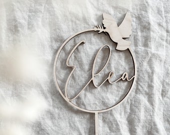 May include: A wooden cake topper with the name "Ela" written in cursive inside a circle. A dove with an olive branch is perched on the top of the circle.
