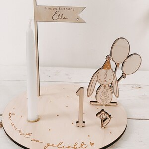 May include: Wooden birthday decoration with a bunny holding balloons, a candle, and a flag that says "Happy Birthday Ella". The base of the decoration has the text "du wirst so geliebt" which translates to "you are so loved".