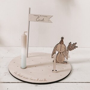 May include: Wooden birthday cake topper with a bunny holding a sprig of leaves, a number one, and a flag that says "Happy Birthday Emil". The base of the topper is a round wooden disc with the text "Du bist ein Geschenk" engraved on it.