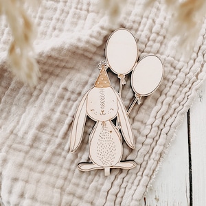 May include: Wooden bunny figurine wearing a party hat and holding two balloons. The bunny has etched details and is set against a neutral-toned fabric background. The balloons are blank, ready for customization.