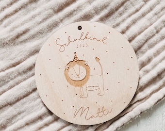 May include: A wooden round ornament with a lion wearing a party hat and the text "Schulkind 2023" and "Matti" engraved on it.