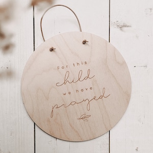 Könnte beinhalten: Ein rundes Holzschild mit einem braunen Lederband zum Aufhängen. Das Schild trägt den Text "For this child we have prayed" in einer kursiv geschriebenen Schrift mit einem kleinen Blattdesign unter dem Text.
