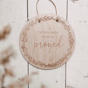 Könnte beinhalten: Ein rundes Holzschild mit einem floralen Kranzdesign und dem Text "Für dieses Kind haben wir gebetet".