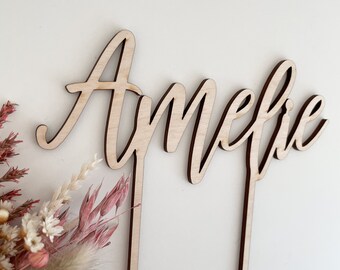 Op de afbeelding: Een houten taarttopper met de naam "Amelie" in cursief schrift. De topper staat op een witte achtergrond met gedroogde bloemen.