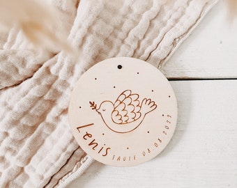 May include: A round wooden ornament with a dove illustration and the name "Lenis" engraved. The dove holds an olive branch. The date "08.08.2022" is also engraved. The ornament has a small hole for hanging.