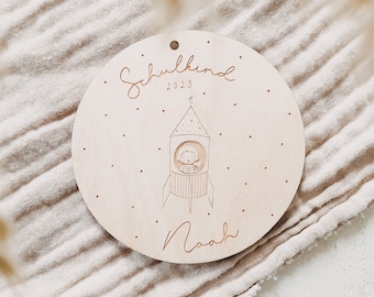 May include: A round wooden plaque with a rocket ship design and the text "Schulkind 2023" and "Noah".