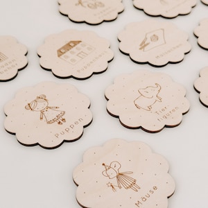 May include: Nine wooden flower-shaped tags with different illustrations and German text. The illustrations include a house, a doll, a mouse, an elephant, and a bicycle. The text includes "Puppenhaus", "Puppen", "Mause", "Tierfiguren", and "Fahrrad".