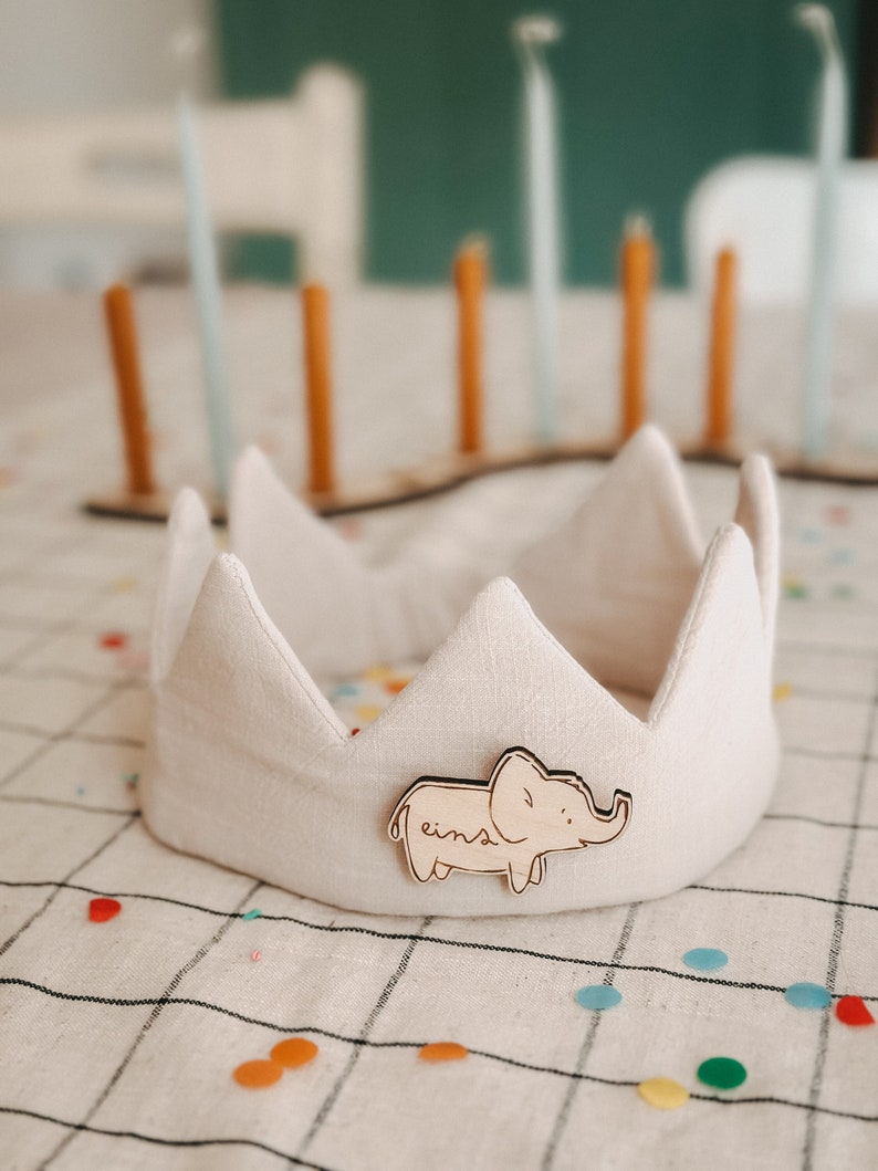 Op de afbeelding: Een witte stoffen kroon met een houten olifant-uitsparing die aan de voorkant is bevestigd. De olifant is bruin geverfd en heeft het woord "eins" erop geschreven. De kroon ligt op een witte tafelkleed met kleurrijk confetti.