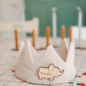 Op de afbeelding: Een witte stoffen kroon met een houten olifant-uitsparing die aan de voorkant is bevestigd. De olifant is lichtbruin en heeft het woord "eins" erop geschreven.