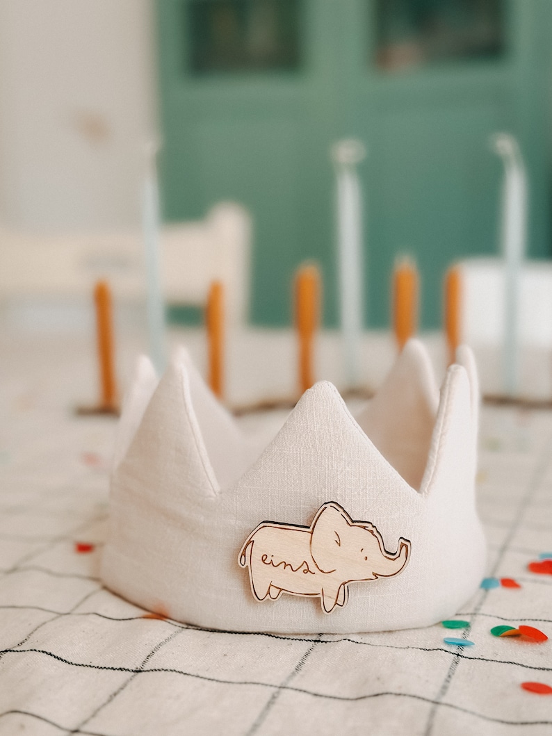 Op de afbeelding: Een witte stoffen kroon met een houten olifant-uitsparing die aan de voorkant is bevestigd. De olifant is lichtbruin en heeft het woord "eins" erop geschreven.
