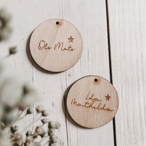 Könnte beinhalten: Zwei runde Holzornamente mit einem Stern und einem Namen, der auf jedes Ornament graviert ist. Das obere Ornament sagt "Ole Mats" und das untere Ornament sagt "Ida Mathilda".