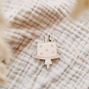 Könnte beinhalten: Ein Holz-Kuchentopper mit weißem Frosting-Design und einer Biene oben drauf. Der Kuchentopper liegt auf einem hellbraunen Stoffhintergrund.