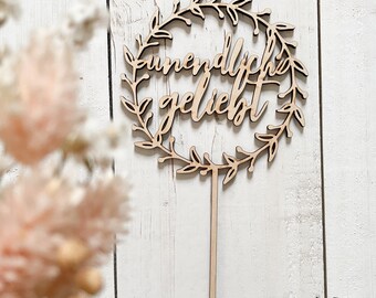 May include: A wooden cake topper with a wreath design and the text "unendlich geliebt" in a cursive font.