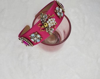May include: A pink headband with a gold bee and pearl embellishments. The headband is decorated with rhinestones and is sitting on a white surface.