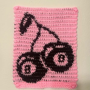 May include: A pink crocheted wall hanging featuring a dark brown cherry design. The edges are finished with a pink border and adorned with small pink heart-shaped embellishments. The handmade piece is likely for home decor.