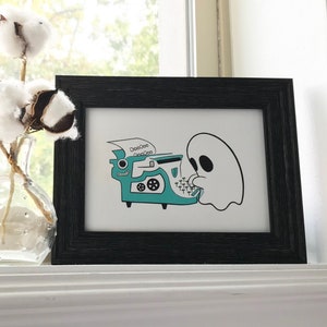 May include: A black framed print with a white background featuring a ghost typing on a teal typewriter with the text "OoooOoo OoooOoo" above it.