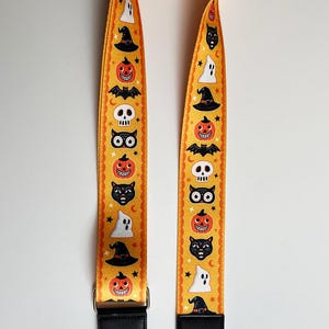 K&ouml;nnte beinhalten: Ein gelber Halloween-Taschenriemen mit sich wiederholenden Mustern aus K&uuml;rbissen, Geistern, Hexenh&uuml;ten, Flederm&auml;usen, Totenk&ouml;pfen und schwarzen Katzen. Der Riemen hat schwarze Lederenden mit silberfarbenen Metallverschl&uuml;ssen.