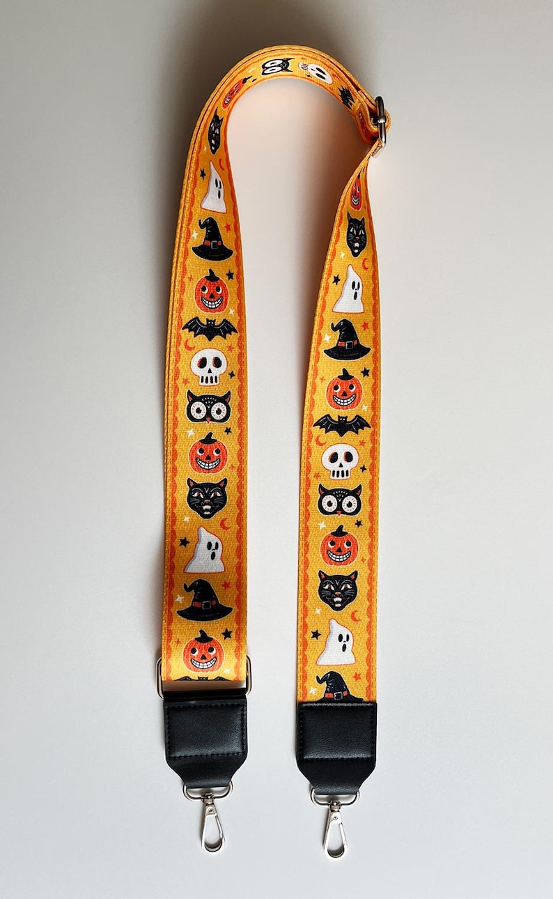 K&ouml;nnte beinhalten: Ein gelber Halloween-Taschenriemen mit sich wiederholenden Mustern aus K&uuml;rbissen, Geistern, Hexenh&uuml;ten, Flederm&auml;usen, Totenk&ouml;pfen und schwarzen Katzen. Der Riemen hat schwarze Lederenden mit silberfarbenen Metallverschl&uuml;ssen.