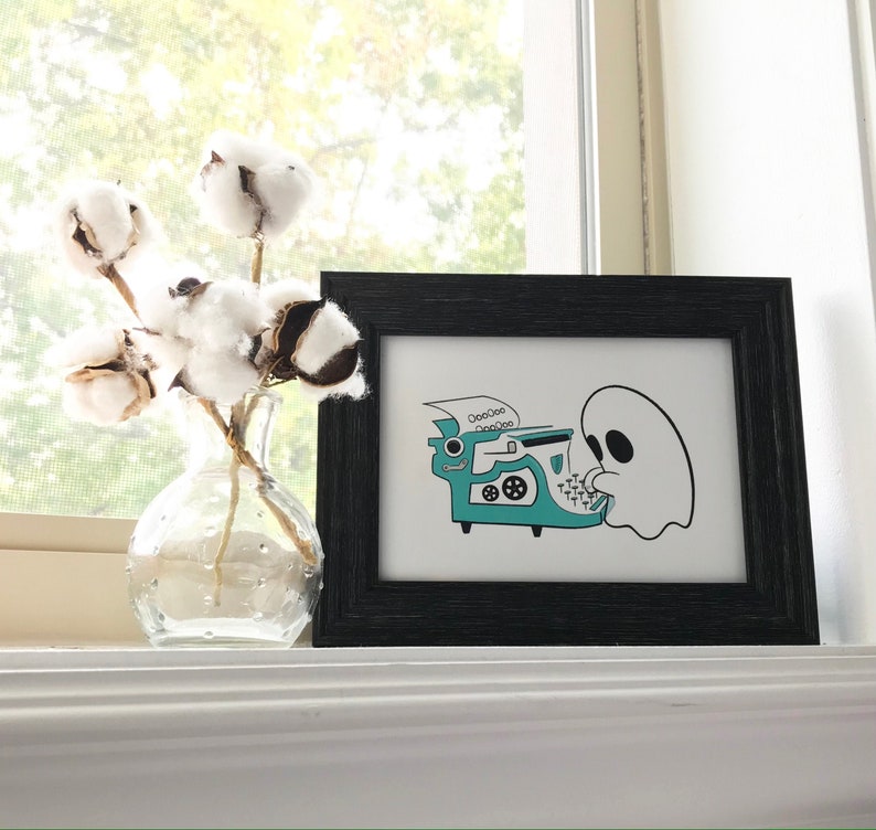 May include: A black framed print with a white background featuring a ghost typing on a teal typewriter with the text "OoooOoo OoooOoo" above it.