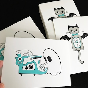 May include: White cards with whimsical illustrations. One card features a ghost typing on a teal typewriter. Other cards show a cartoon bat with a bow tie and a bat holding a clock. The cards are stacked on a black surface.