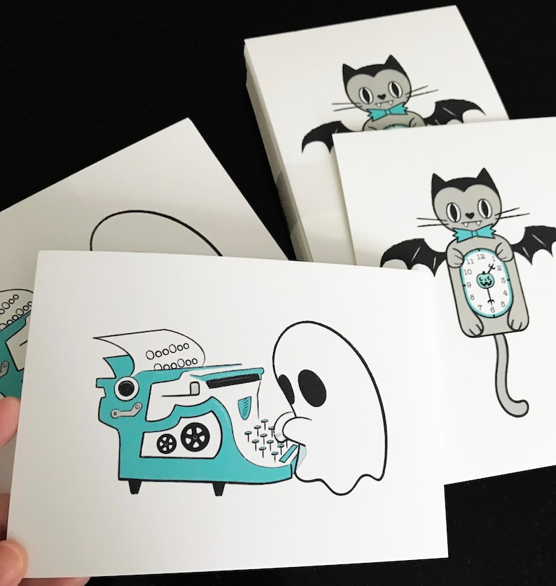 May include: White cards with whimsical illustrations. One card features a ghost typing on a teal typewriter. Other cards show a cartoon bat with a bow tie and a bat holding a clock. The cards are stacked on a black surface.