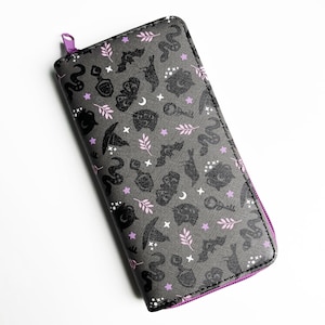 May include: A rectangular zip-around wallet in dark gray featuring a repeating pattern of black silhouettes including snakes, bats, cats, witch hats, keys, and potion bottles, accented with small light pink leaves and white stars.