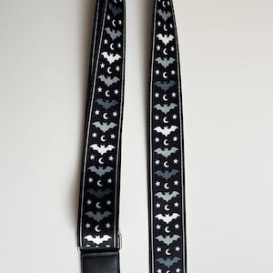 May include: Black and white patterned bag strap featuring a repeating design of bats, stars, and crescent moons. The strap has black leather ends with silver-tone metal hardware. The strap is designed for a bag.