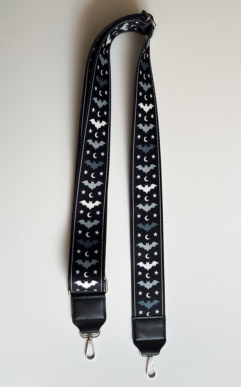 May include: Black and white patterned bag strap featuring a repeating design of bats, stars, and crescent moons. The strap has black leather ends with silver-tone metal hardware. The strap is designed for a bag.