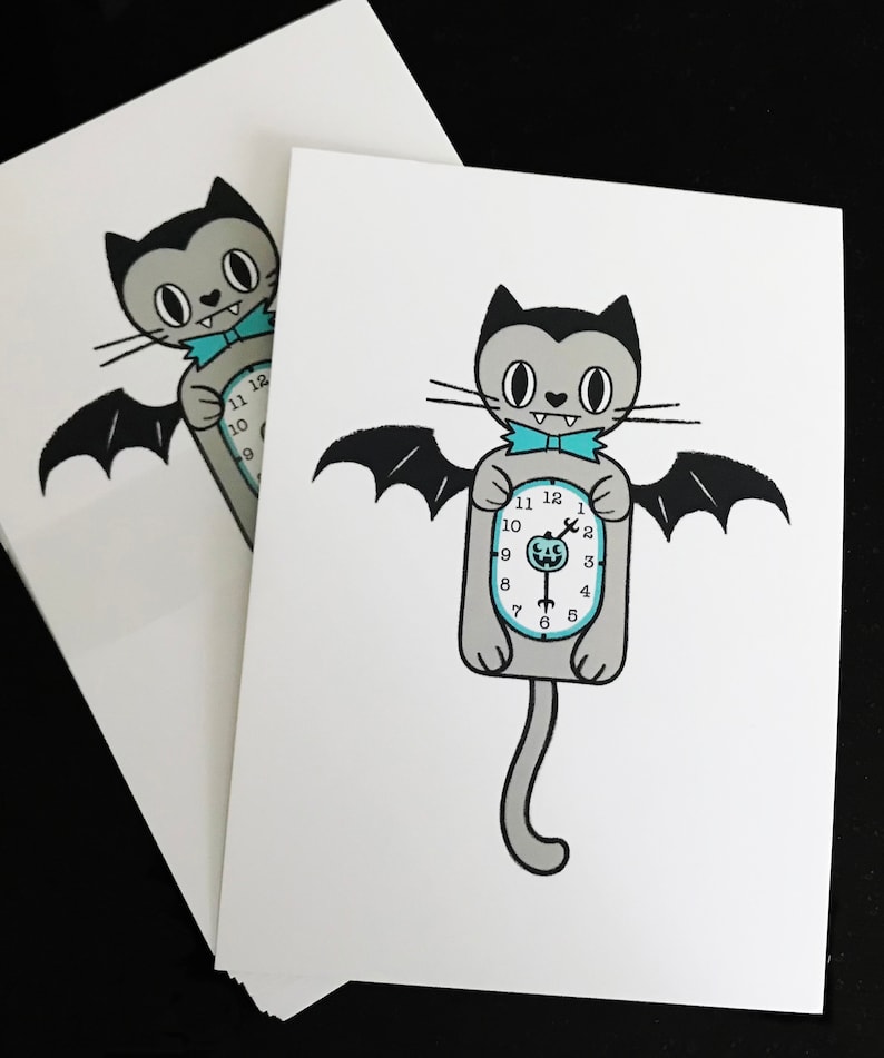 May include: Two white cards featuring a cartoon cat with bat wings and a clock body. The cat is gray with black accents, a blue bow tie, and a clock face showing the time. The cards are stacked on a black surface.
