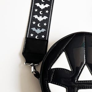 May include: Black, glossy, round handbag shaped like a jack-o'-lantern with white accents. The strap is black with a pattern of bats, moons, and stars. A silver clasp connects the strap to the bag.