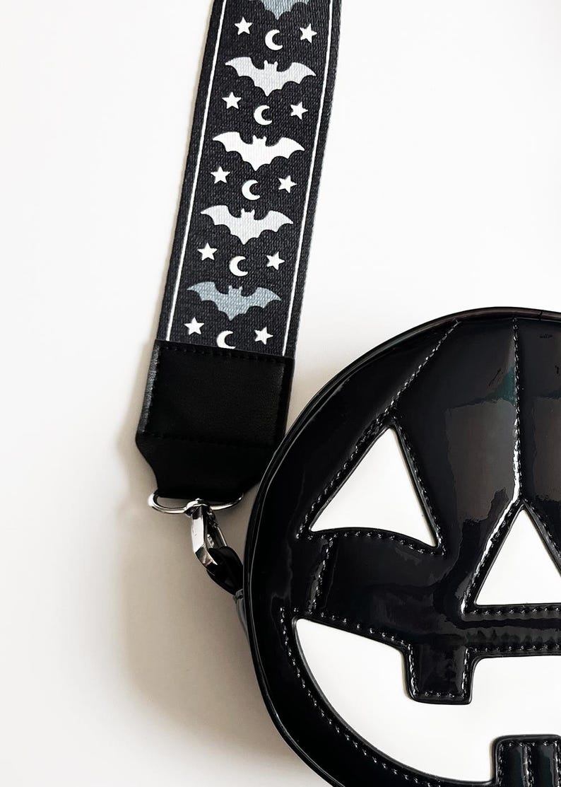 May include: Black, glossy, round handbag shaped like a jack-o'-lantern with white accents. The strap is black with a pattern of bats, moons, and stars. A silver clasp connects the strap to the bag.