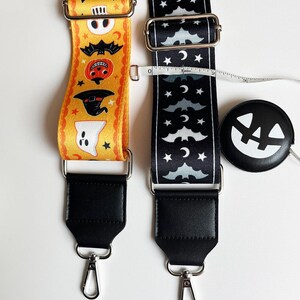 May include: Two Halloween-themed bag straps. One is yellow with orange and black Halloween characters. The other is black with white bats, moons, and stars. Both straps have black leather ends and silver metal hardware. A black and white pumpkin-shaped tape measure is also present.