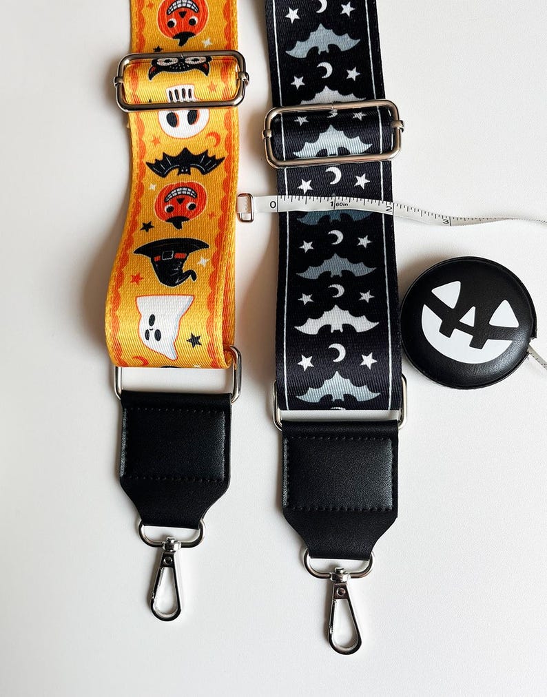 May include: Two Halloween-themed bag straps. One is yellow with orange and black Halloween characters. The other is black with white bats, moons, and stars. Both straps have black leather ends and silver metal hardware. A black and white pumpkin-shaped tape measure is also present.